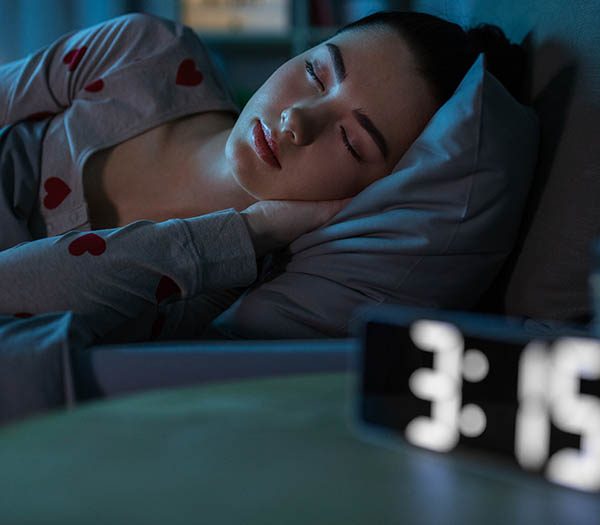 teenage girl sleeping in bed next to alarm clock showing 3:15 a.m.