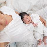 tired mother suffering from postnatal depression sitting on bed holding her sleeping baby, parenting stress